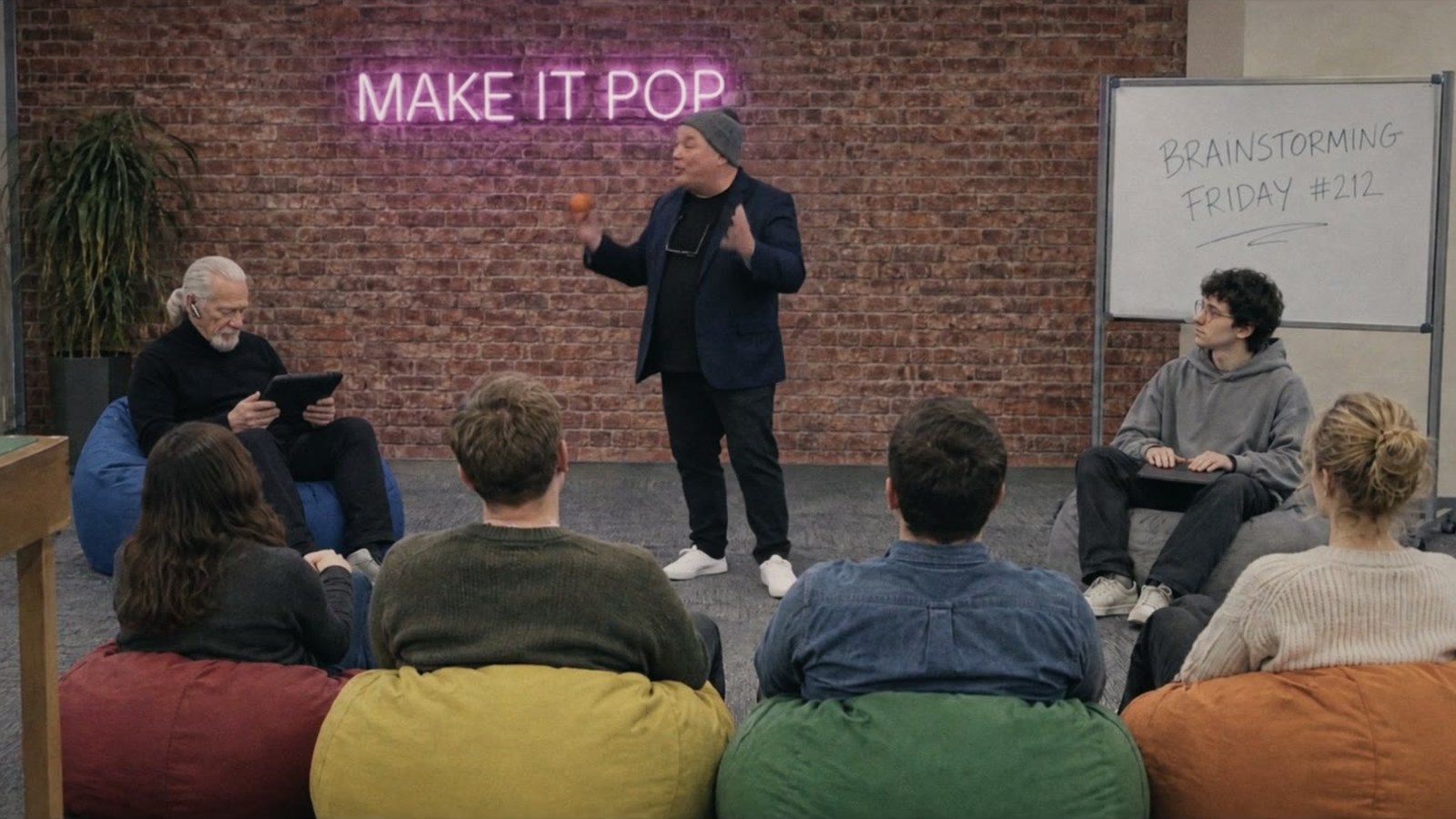 Großraumbüro mit Neonschrift „Make it pop“: Ein Agenturchef steht vor seinem Team, das auf Sitzsäcken sitzt.